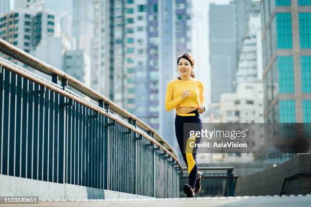 smiling young asian sports woman jogging on city bridge outdoors against urban city skyline - commercial buildings hong kong morning stock pictures, royalty-free photos & images