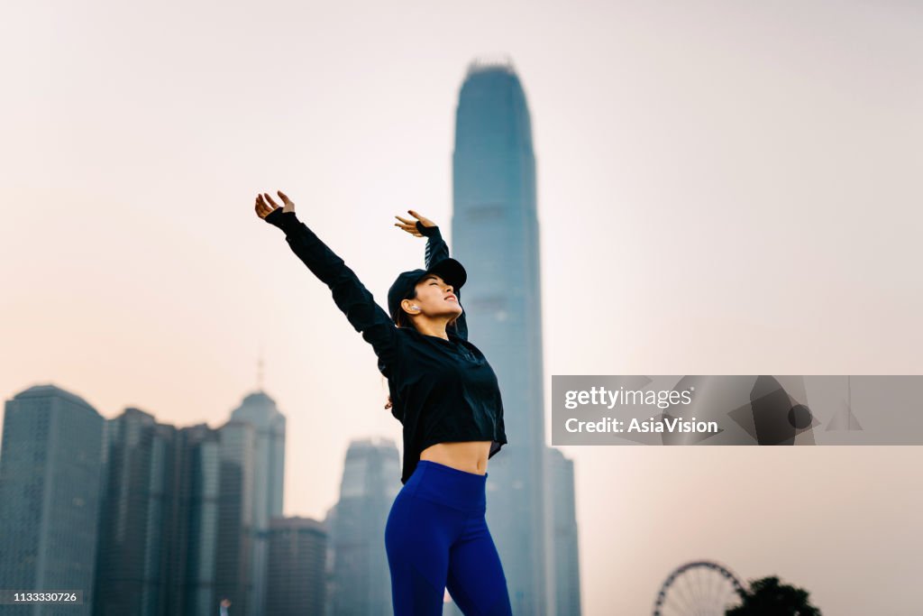 Confidence and smiling Asian sports woman stretching arms overhead outdoors against urban cityscape at sunset