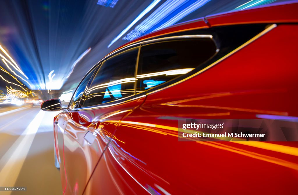 Red electric powered car drives on city highway while night - streaking street lights and signs.