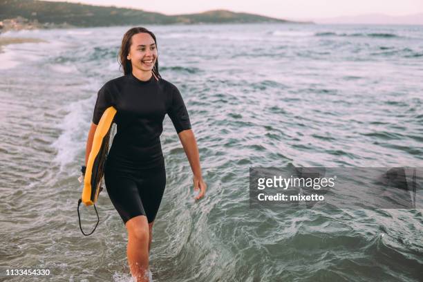 surfer meisje lopen door het water - surfer wetsuit stockfoto's en -beelden