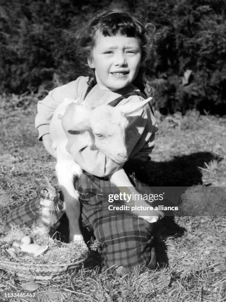 Ein kleines Mädchen sitzt mit einem Osterlämmchen im Arm auf einer Wiese, vor ihr ein Osternest mit Ostereiern und einem Osterhasen aus Schokolade....