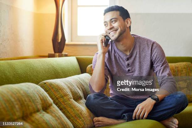 smiling young man answering smart phone on couch - answering stock pictures, royalty-free photos & images