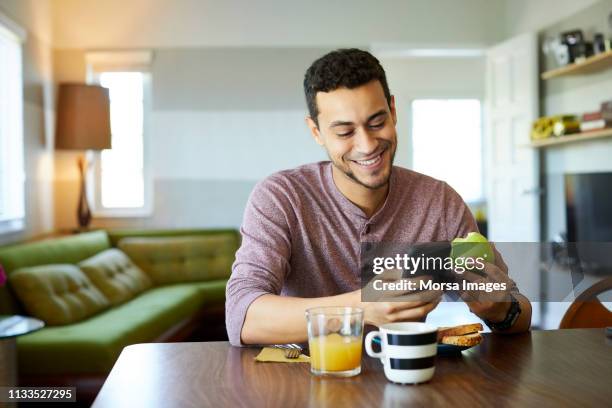 lächelnde mann mit handy, während er apfel hält - mobile device on table stock-fotos und bilder