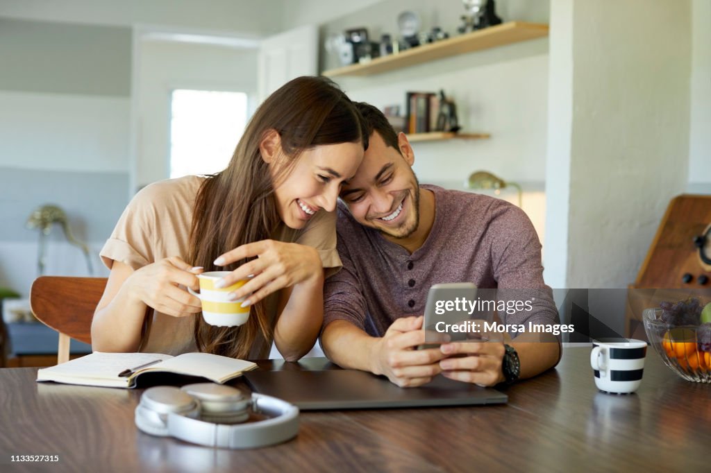 Pareja alegre utilizando el teléfono móvil en el comedor
