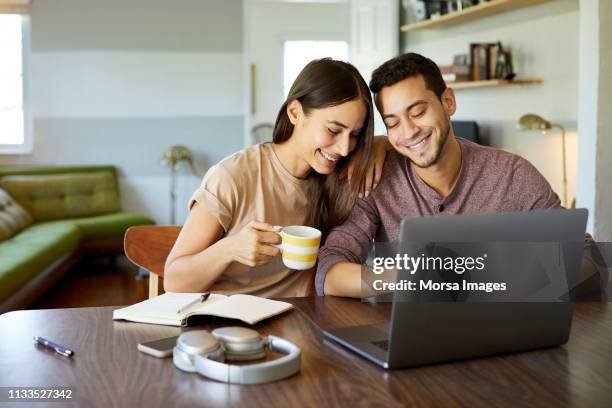 smiling young couple using laptop on table at home - hispanic couple stock pictures, royalty-free photos & images