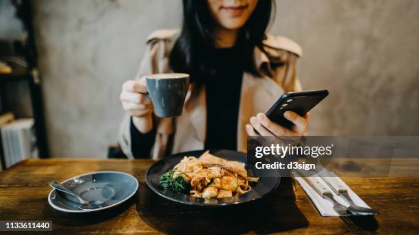 smiling young woman checking on smartphone while having coffee during lunch break in a restaurant - dinner program stock pictures, royalty-free photos & images