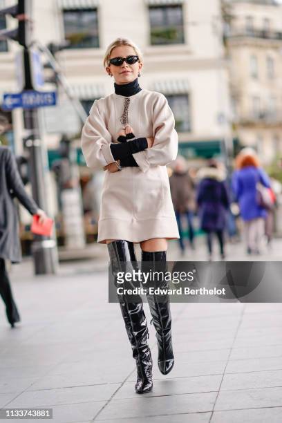 Caroline Daur wears sunglasses, a black turtleneck, a beige dress with pompons at the collar, shiny black patent leather thigh high boots, outside...