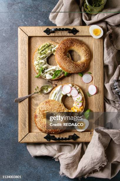 Variety of homemade bagels with sesame seeds, cream cheese, pesto sauce, eggs, radish, herbs served on wooden tray with cloth and ingredients above...