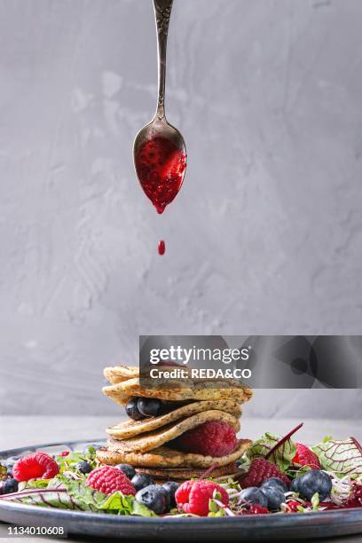 Vegan chickpea pancakes served in plate with green salad young beetroot leaves, sprouts, berries, berry sauce flowing from spoon over grey kitchen...