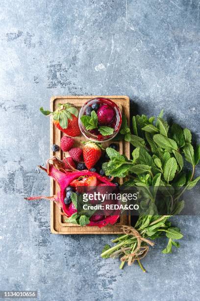Vegan fruit salad with berries and mint served in pink dragon fruit with ingredients above on wooden serving board over blue texture background Top...