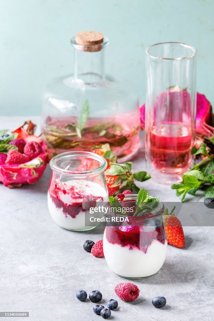 Jars of natural white yogurt with berry sauce, fruit salad with pink dragon fruit, berries and mint, served with bottle of lemonade on grey table Healthy eating