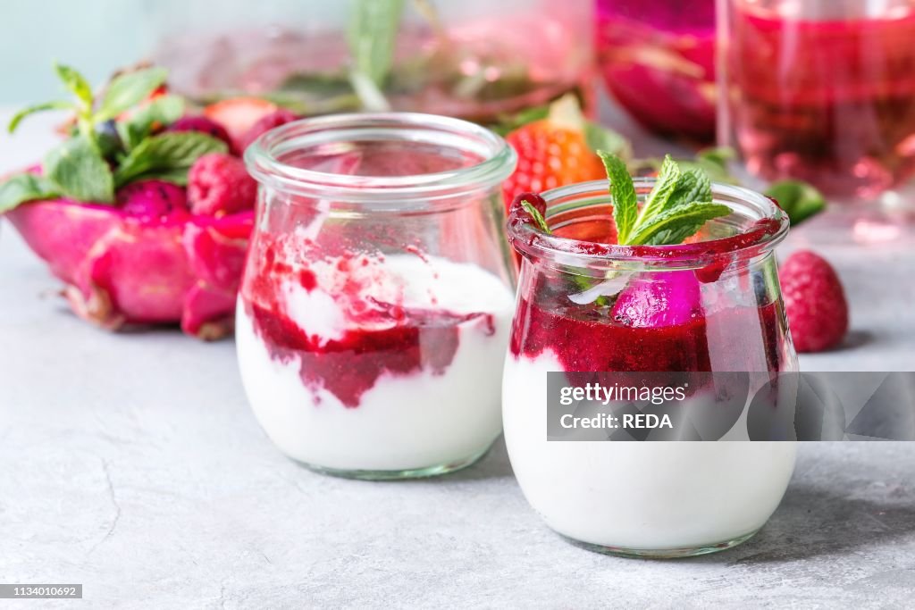 Jars of natural white yogurt with berry sauce, fruit salad with pink dragon fruit, berries and mint, served with bottle of lemonade on grey table Close up Healthy eating
