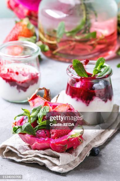 Vegan fruit salad with berries and mint served in pink dragon fruit on cloth with jars of yogurt, bottle of lemonade, ingredients above on grey table...