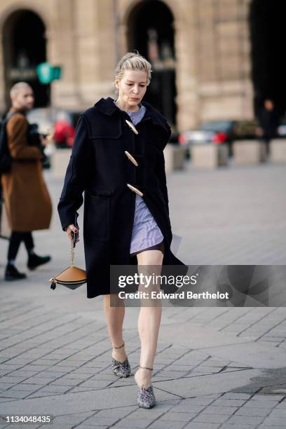 Guest wears a black coat with brass fasteners, a brown and black cone shaped bag, grey python pattern pumps, outside Louis Vuitton, during Paris...