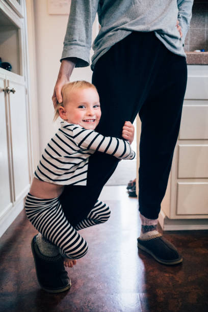 young girl (age 3 years) clinging playfully to her mother's leg in the family home. - kids hanging on mom stock pictures, royalty-free photos & images