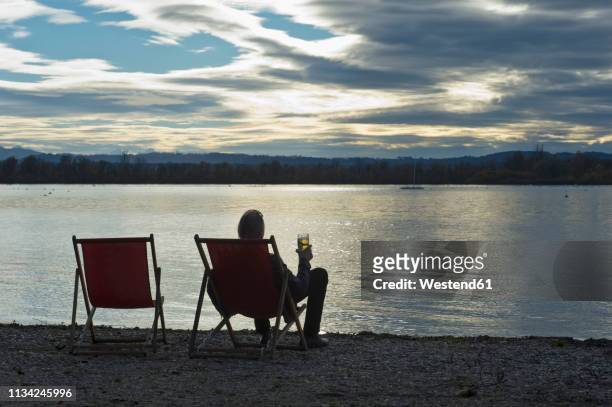 germany, man sitting at lakeshore of ammersee with glass of beer watching sunset - ammersee stockfoto's en -beelden