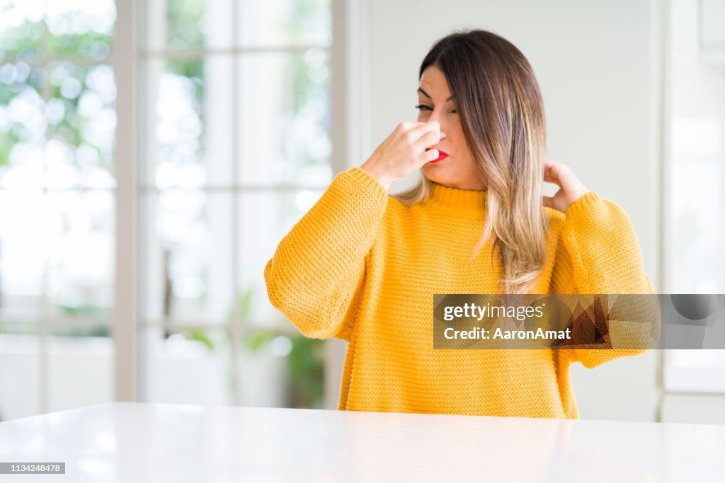 Young beautiful woman wearing winter sweater at home smelling something stinky and disgusting, intolerable smell, holding breath with fingers on nose. Bad smells concept.