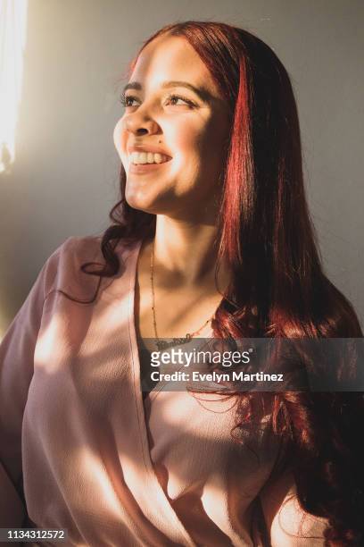 Young Latina woman looking to the side, smiling. Woman has dyed red hair and is wearing a necklace and pink top.