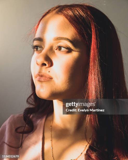 Portrait of young Latina woman looking off to the side, not smiling. Young woman has red hair and a mole near her mouth. Sunlight is on her face.