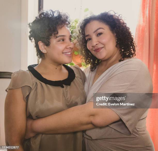 Mother hugging daughter by a bedroom window. Woman has arms wrapped around young woman. House plant and curtains in the background.