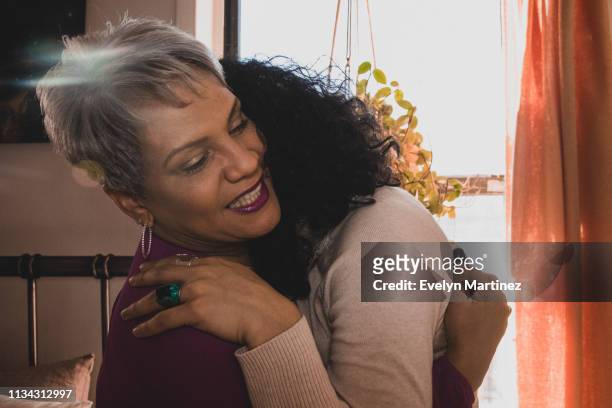 Latina mother with gray hair embracing her daughter with black hair. House plant, curtains and bed in the background.