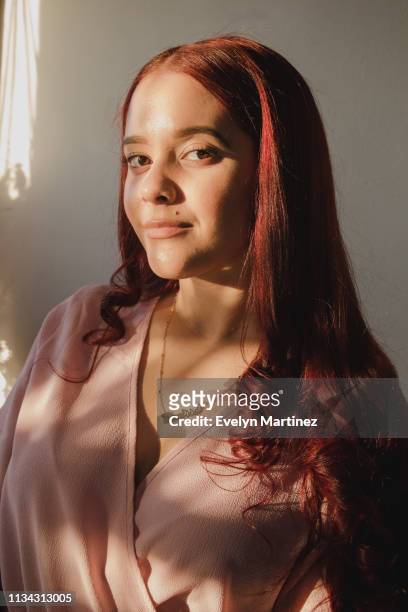 Portrait of Latina woman with red hair standing in the sunlight. Woman has a mole, is wearing a necklace and a pink top.