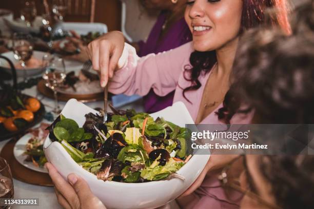 Latina women passing salad serving bowl at the dinner table. Woman with red hair serves herself salad. Woman next to her is holding the ceramic bowl.