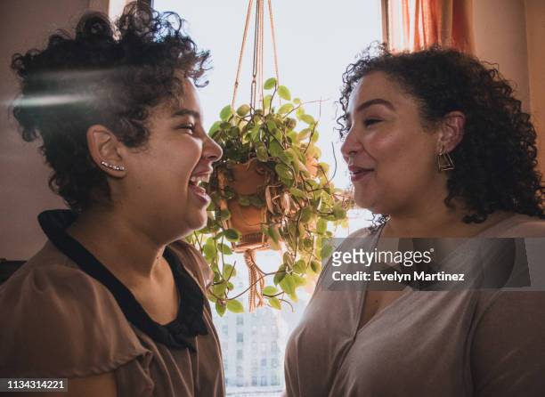 Latina daughter laughing and Latina mother smiling. A house plant and curtain are in the background.