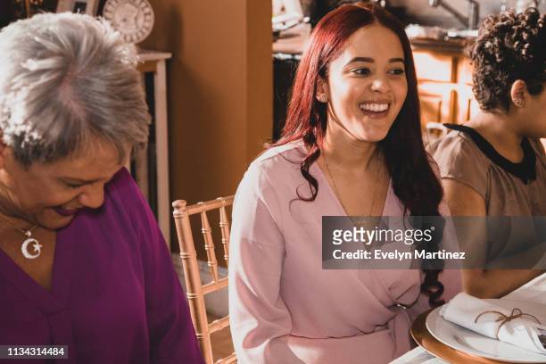 Candid laughter at dinner table. Latina woman with short gray hair mid laugh sitting next to Latina woman with long red hair. Both are looking away from the camera. Partial view of young woman on the right side of the photo.