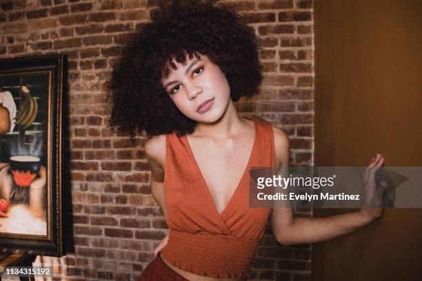 Young Latina wearing orange top and orange bottom standing with one hand on her waist, one hand on the wall. Girl has a short afro. Background is an exposed brick wall and a painting.