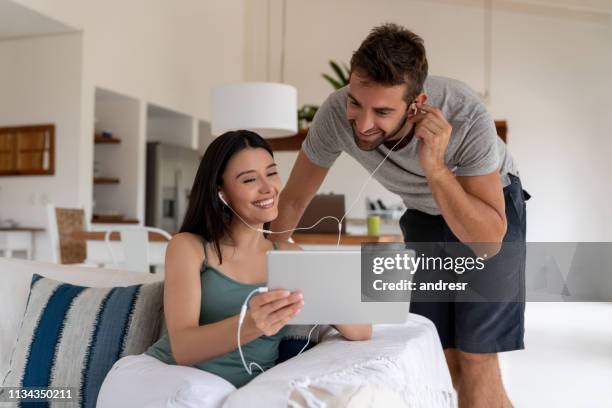 happy couple at home watching videos on a tablet computer and sharing headphones - sharing headphones stock pictures, royalty-free photos & images