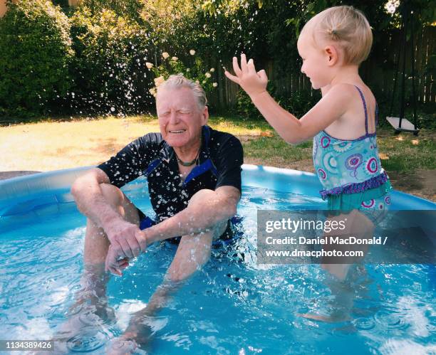 an elderly grandfather gets splashed by his granddaughter (age 3 years) while sitting in a wading pool in their family yard on a hot summer day. - old and young com stockfoto's en -beelden