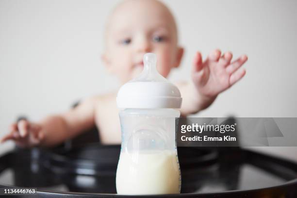 baby boy sitting in high chair reaching for baby bottle - baby bottle stockfoto's en -beelden