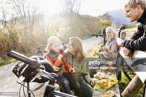 family with bicycles resting on bench by roadside eating picnic smiling - familie unterwegs stock-fotos und bilder