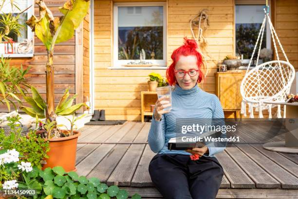 portrait of smiling senior woman with red dyed hair sitting on terrace in front of her house reading a book - anti ageing stockfoto's en -beelden