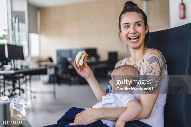 young mother with tattoos sitting in office with her baby on lap, eating a sandwich - millennial generation stock-fotos und bilder