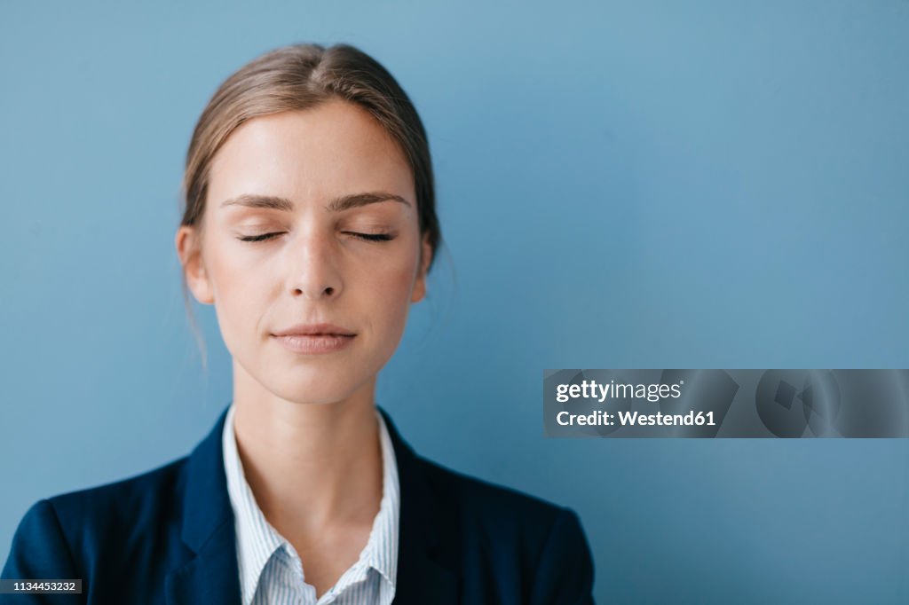 Portrait of a young businesswoman against blue background, relaxing with eyes closed