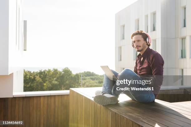 man sitting cross-legged on a rooftop terrace, using laptop, wearing headphones - rooftop pool stock-fotos und bilder