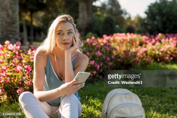 smiling young woman sitting in park with cell phone and earbuds - woman smiling park summer stock-fotos und bilder