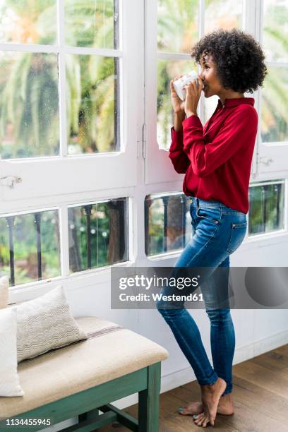 woman looking out of window at home drinking cup of coffee - african american woman barefoot stock pictures, royalty-free photos & images