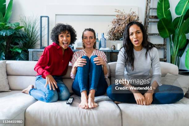 three excited women on couch at home watching tv - stressed young woman sitting on couch stock-fotos und bilder