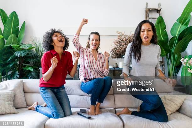 three excited women on couch at home watching tv and cheering - cheering stock pictures, royalty-free photos & images