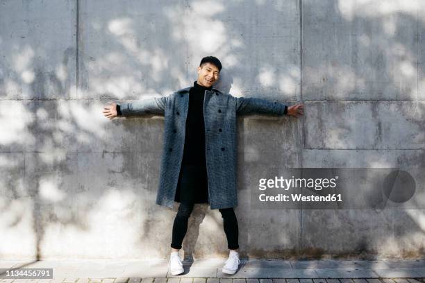 laughing young man wearing turtleneck pullover and grey coat standing in front of concrete wall - mann ganzkörper lachen stehen stock-fotos und bilder