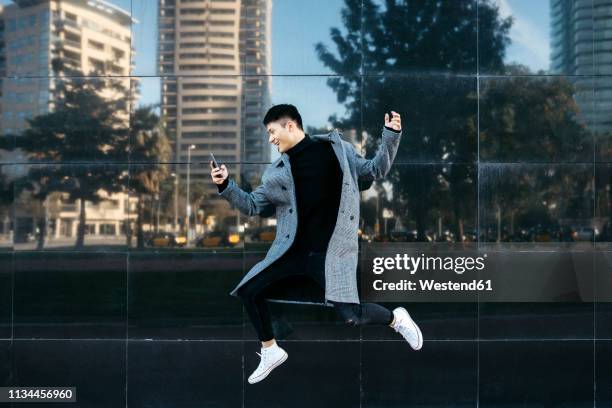 spain, barcelona, happy young man jumping in the air while looking at cell phone - stadt begeisterung stock-fotos und bilder