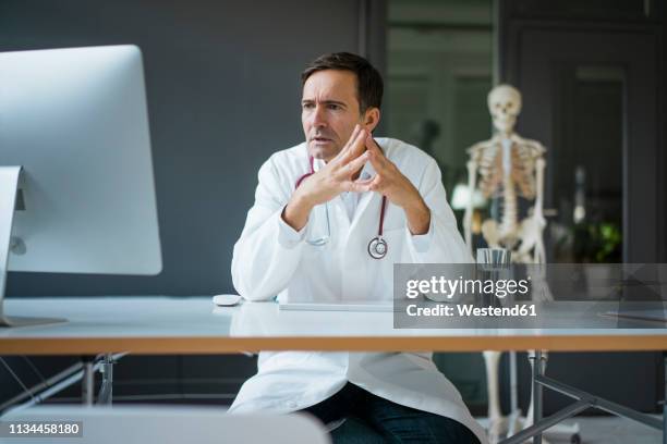 serious doctor sitting at desk in medical practice with skeleton in background - anatomista fotografías e imágenes de stock