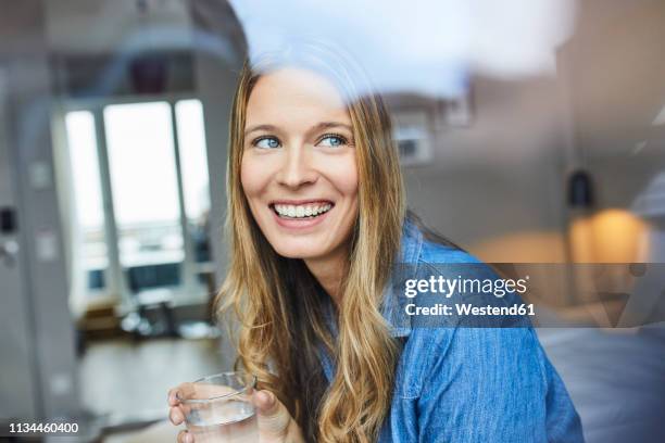 happy young woman looking out of window - drinking water glass woman stock-fotos und bilder