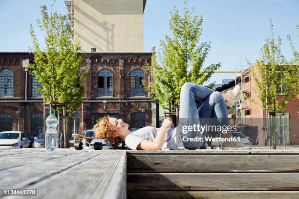 smiling young woman with longboard lying on bench in urban surrounding - woman longboard stock pictures, royalty-free photos & images