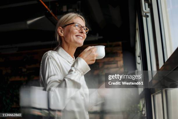 smiling businesswomanholding cup of coffee looking out of window - cofee stock-fotos und bilder