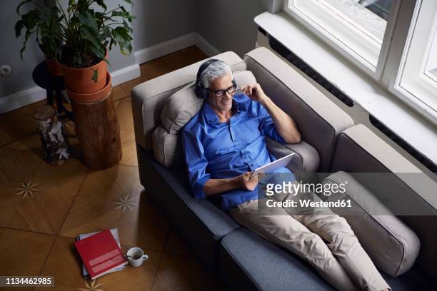 smiling mature man with tablet and headphones relaxing on couch at home - movie still stock-fotos und bilder