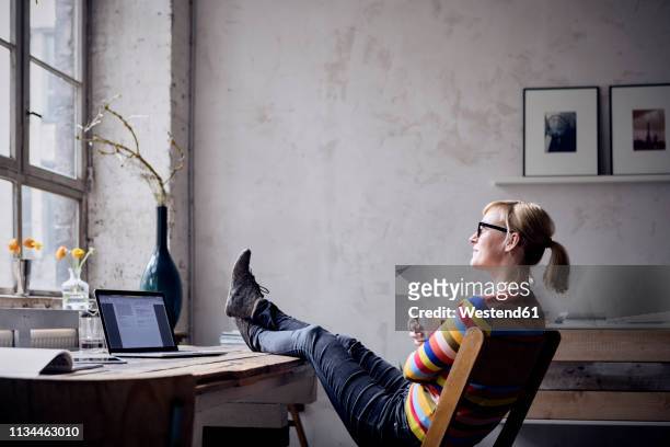 smiling woman sitting with feet up at desk in a loft looking through window - feet up stock pictures, royalty-free photos & images